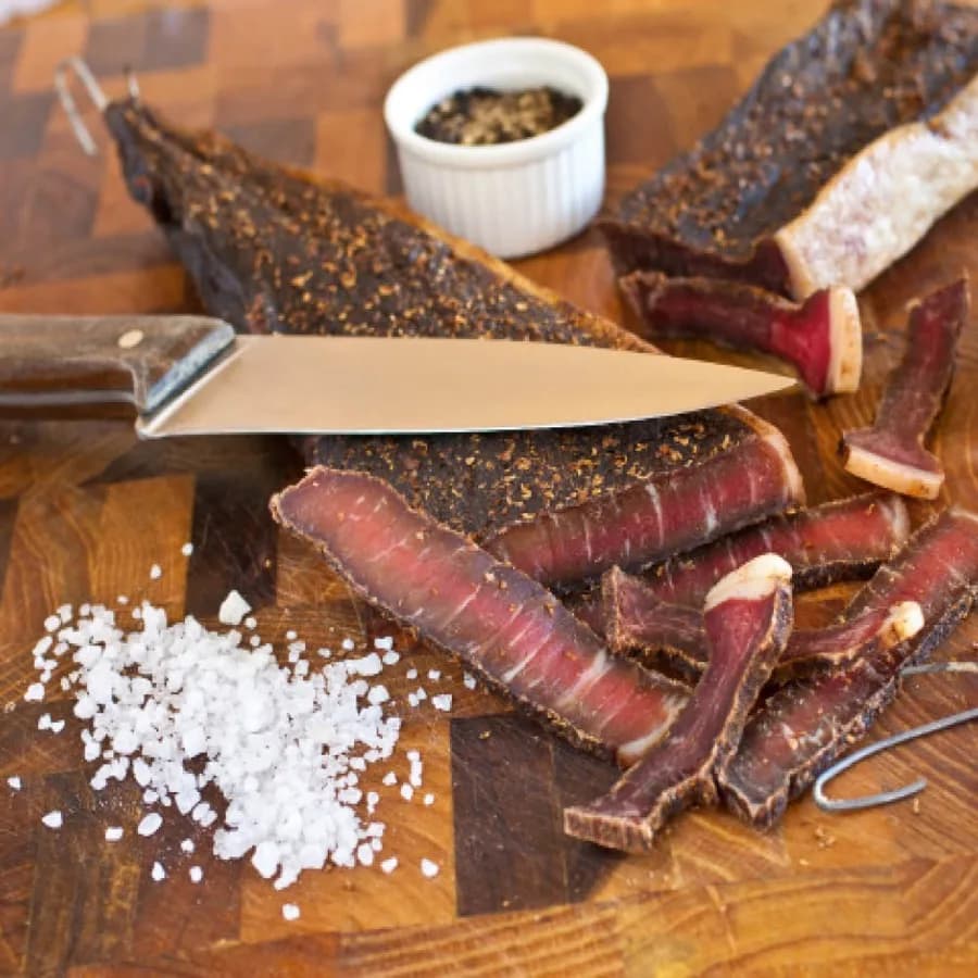 Sliced biltong on a wooden board with salt and a knife